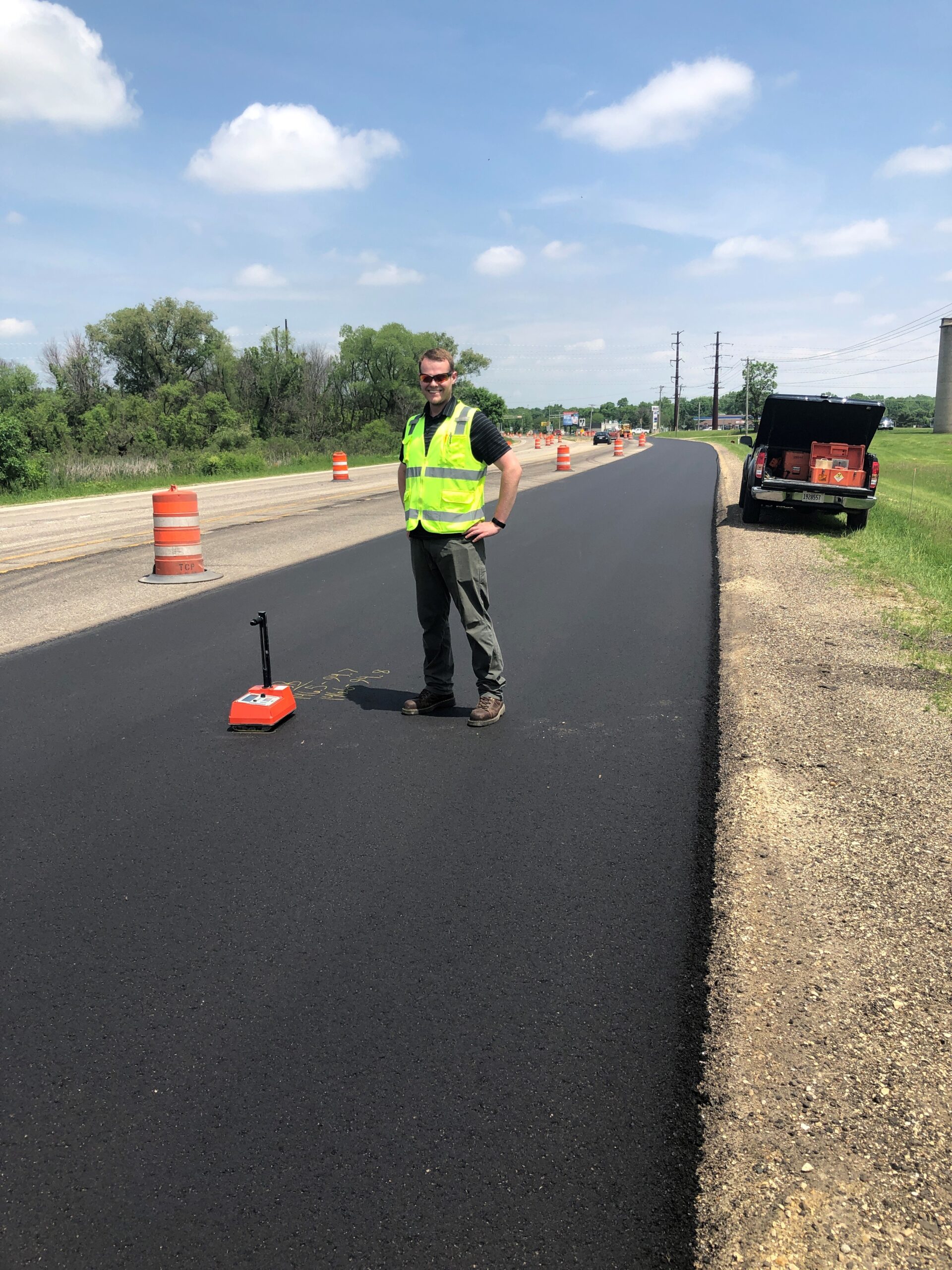 A person in a high-visibility vest stands on a newly paved road, next to measuring equipment. Orange traffic barrels line the road on a sunny day.