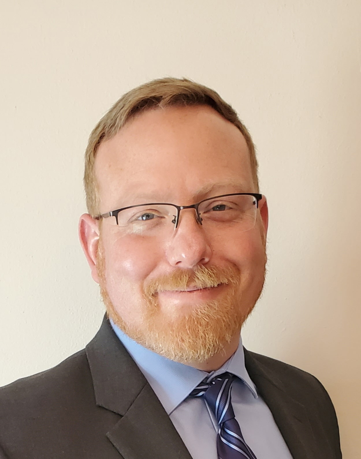 A man with light hair and glasses smiles warmly. He's wearing a dark suit, light blue shirt, and striped tie, set against a plain wall background.