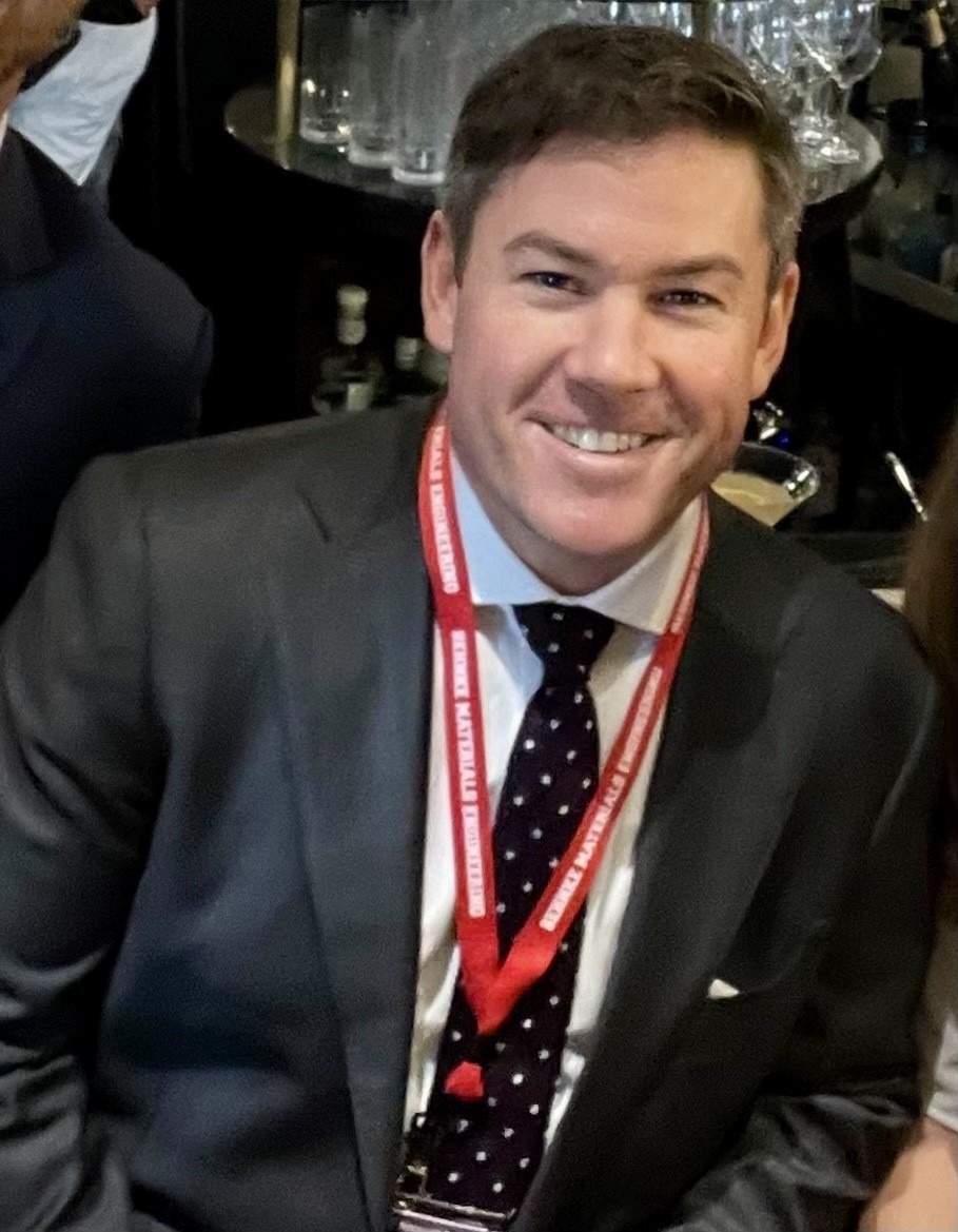 Smiling man in a suit and polka dot tie with a red lanyard, sitting against a backdrop of glassware and dim lighting, conveying sophistication.