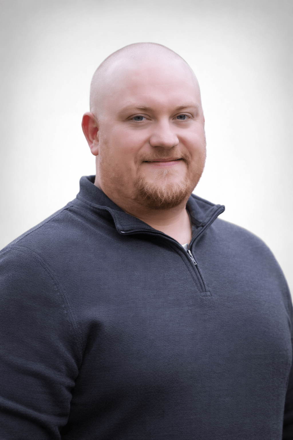Bald man with a trimmed beard smiling slightly, wearing a dark gray zip-up sweater. The background is plain white, creating a calm, approachable tone.